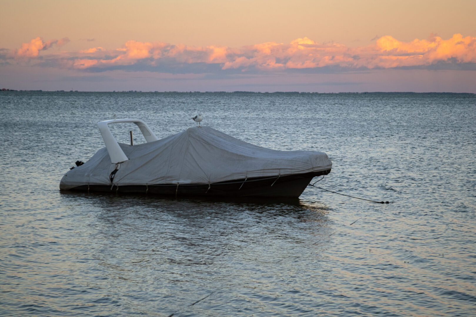 A covered boat is anchored on a calm body of water during sunset, with pink-tinged clouds in the background.