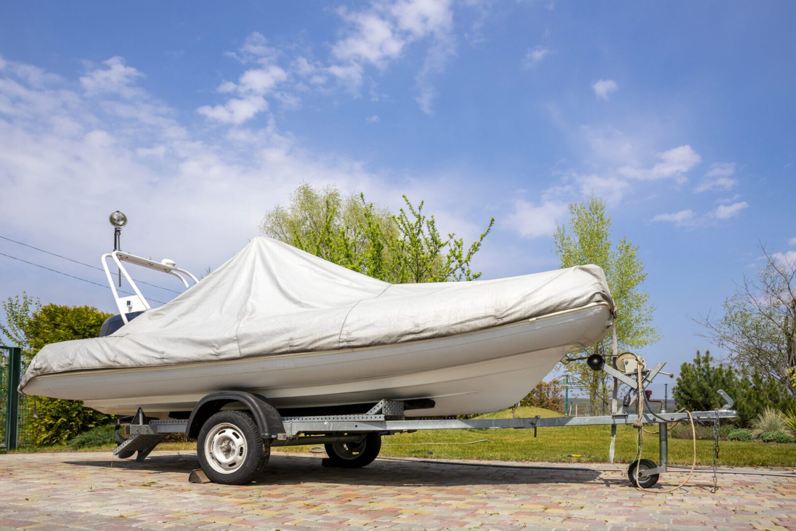 Big modern inflatable motorboat ship covered with grey or white protection tarp standing on steel semi trailer at home backyard on bright sunny day with blue sky on background. Boat vessel storage A white inflatable boat with a protective cover is secured on a trailer, parked on a paved area. Trees and a blue sky are in the background.
