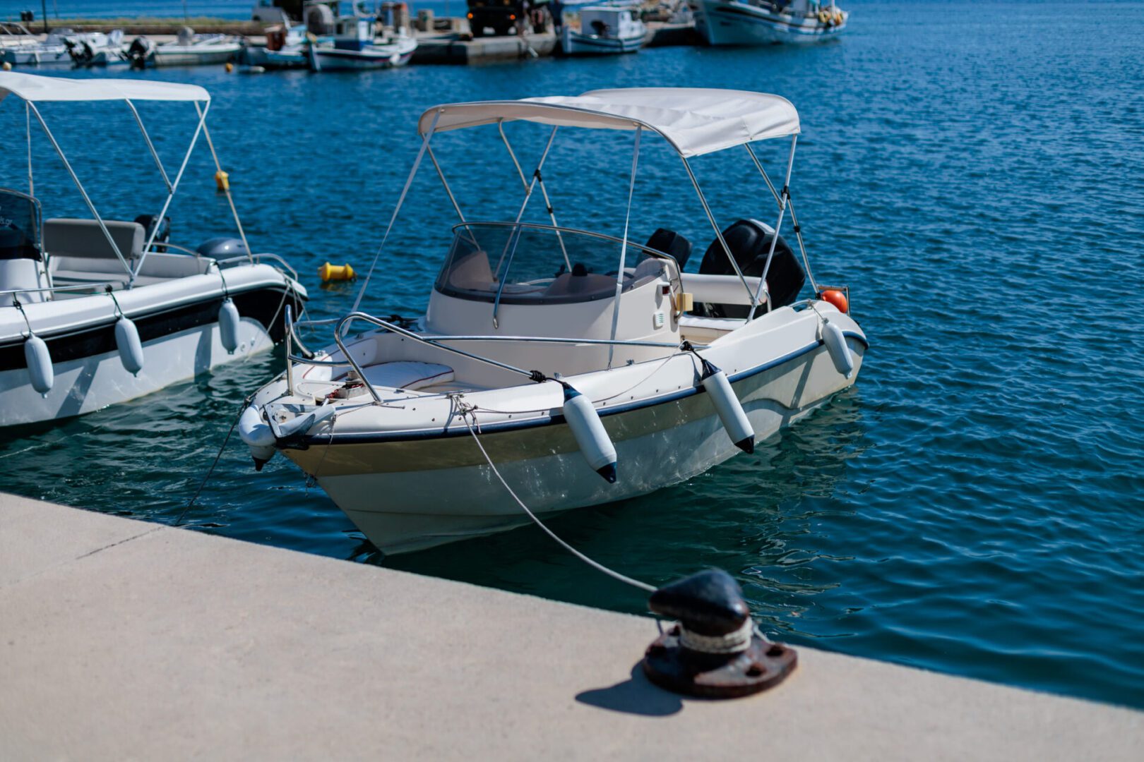 A small motorboat with a canopy is docked at a marina, secured by a rope to a cleat on a concrete pier. Other boats are visible in the background on a sunny day.