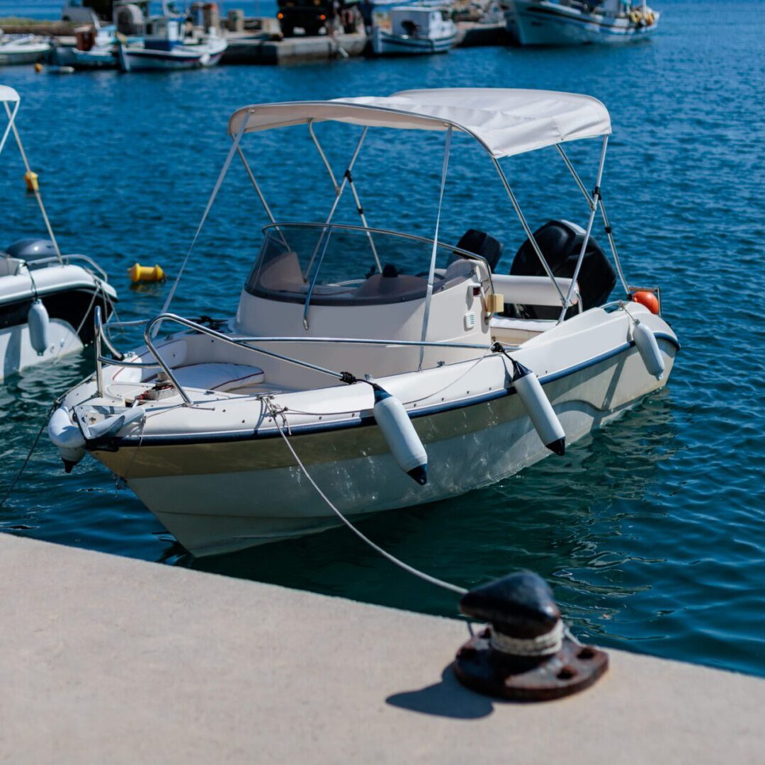 Rental boat docked at a marina on a sunny day A small motorboat with a canopy is docked at a marina, secured by a rope to a cleat on a concrete pier. Other boats are visible in the background on a sunny day.